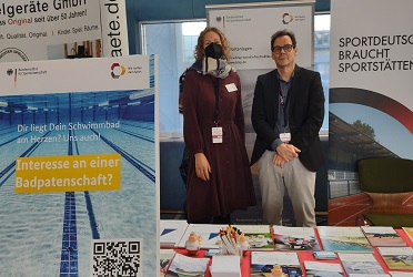 Das Bild zeigt den BISp-Messestand mit Aufstellern auf der sportinfra 2022. Eine Frau und ein Mann stehen hinter dem Tisch, auf dem Prospekte ausgelegt sind (verweist auf: BISp bei der sportinfra zum Thema "Klima­neutrale Sport­stätten")