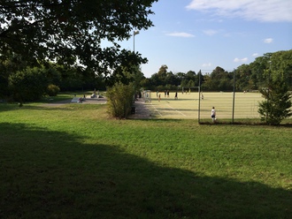 Auf dem Bild ist ein Fußballplatz mit einem Rasenbelag zu sehen. Darauf spielen Menschen Fußball. Daneben ist eine kleine Skateanlage zu sehen.